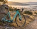 Bicicleta gravel turquesa con manillar curvo, frenos de disco y transmisión de un solo plato, apoyada en un mojón de piedra junto a una pista de tierra entre encinas al amanecer