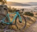 Bicicleta gravel turquesa con manillar curvo, frenos de disco y transmisión de un solo plato, apoyada en un mojón de piedra junto a una pista de tierra entre encinas al amanecer